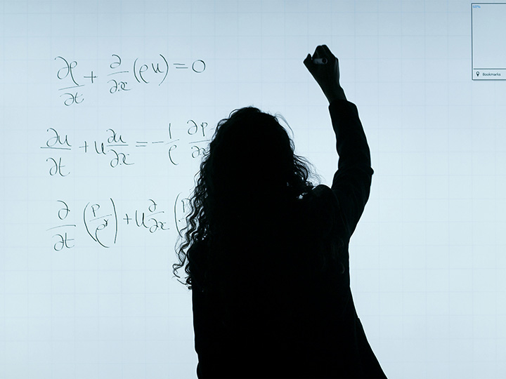 Woman against the light writing mathematical formulae on a whiteboard