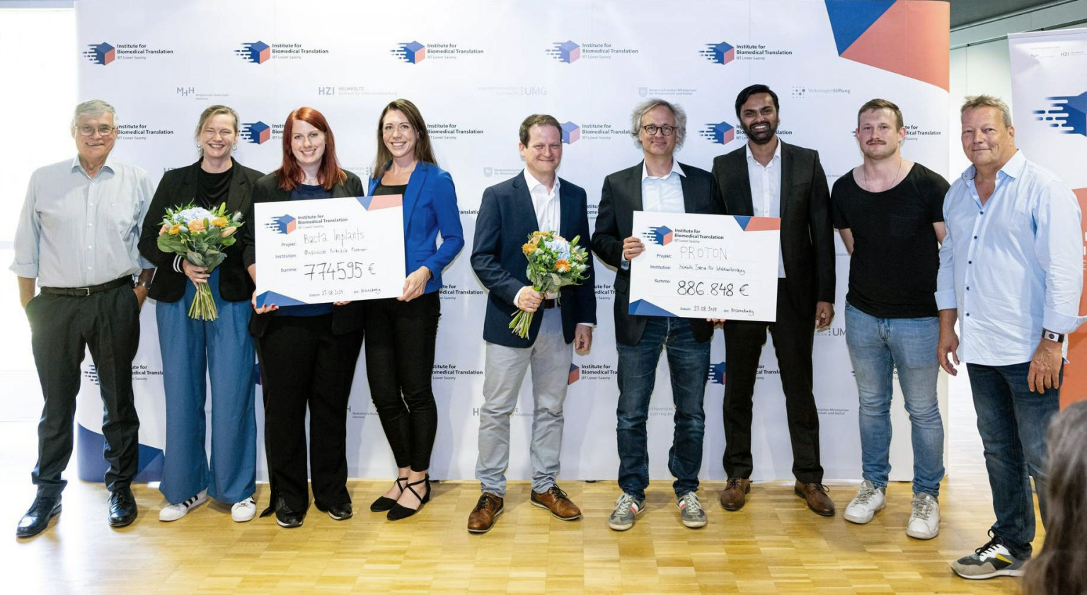 Neun Menschen stehen nebeneinander, zwei halten Blumensträuße, zwei jeweils einen großen Scheck mit Projekttitel und Fördersumme