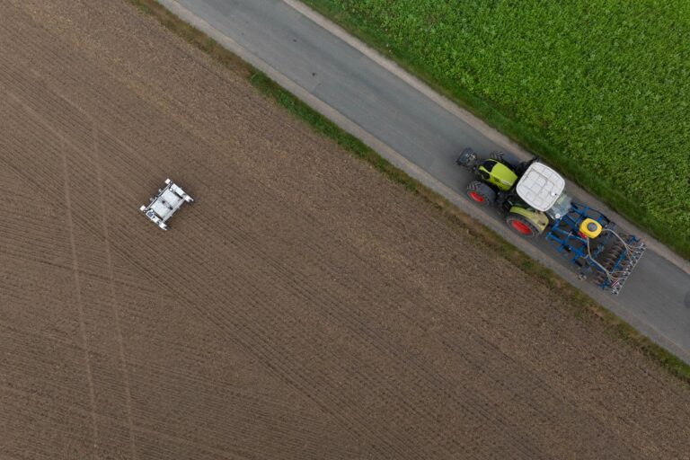 Ein Feld aus der Vogelperspektive mit einem Feldroboter in der linken Mitte neben einem Traktor auf dem Feldweg