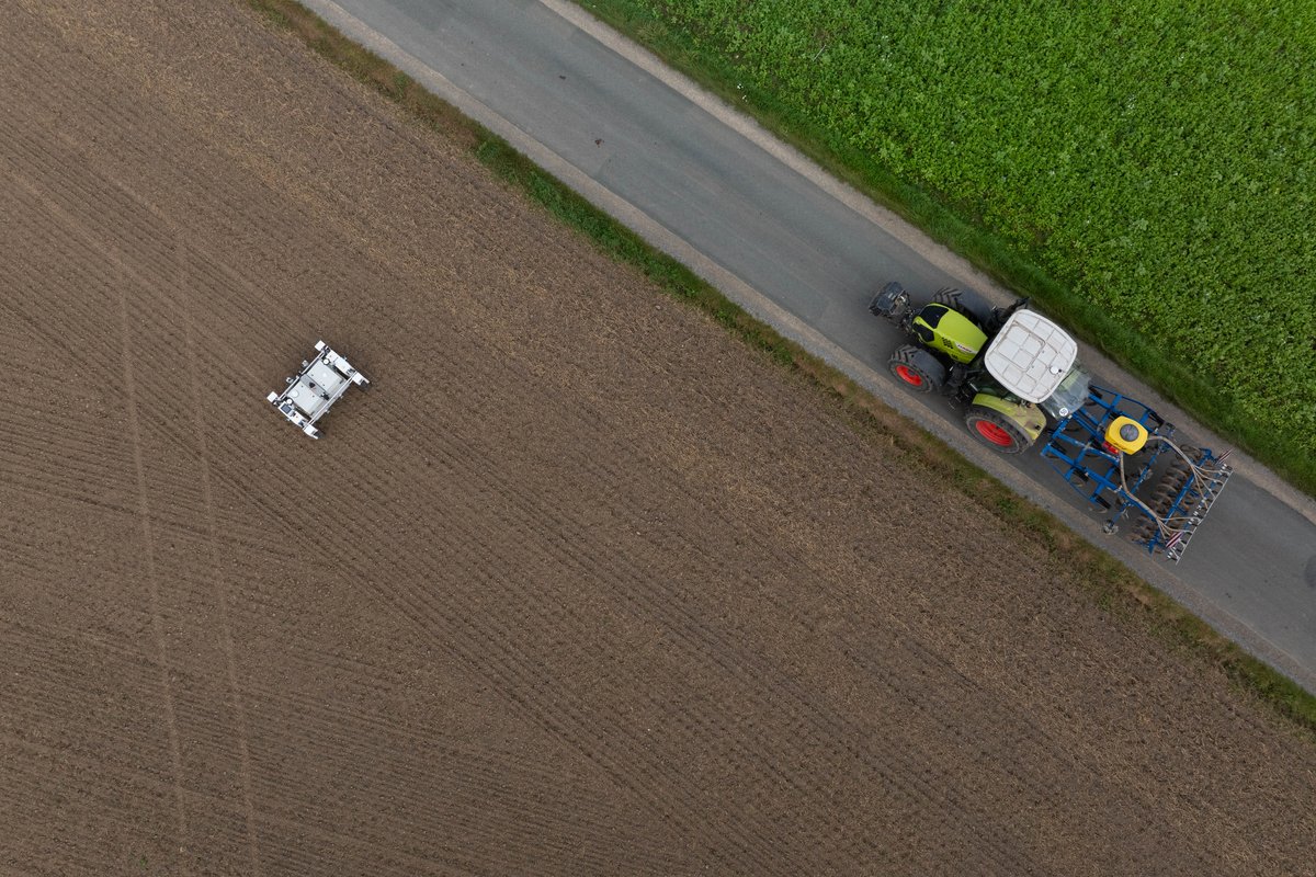 Ein Feld aus der Vogelperspektive mit einem Feldroboter in der linken Mitte neben einem Traktor auf dem Feldweg