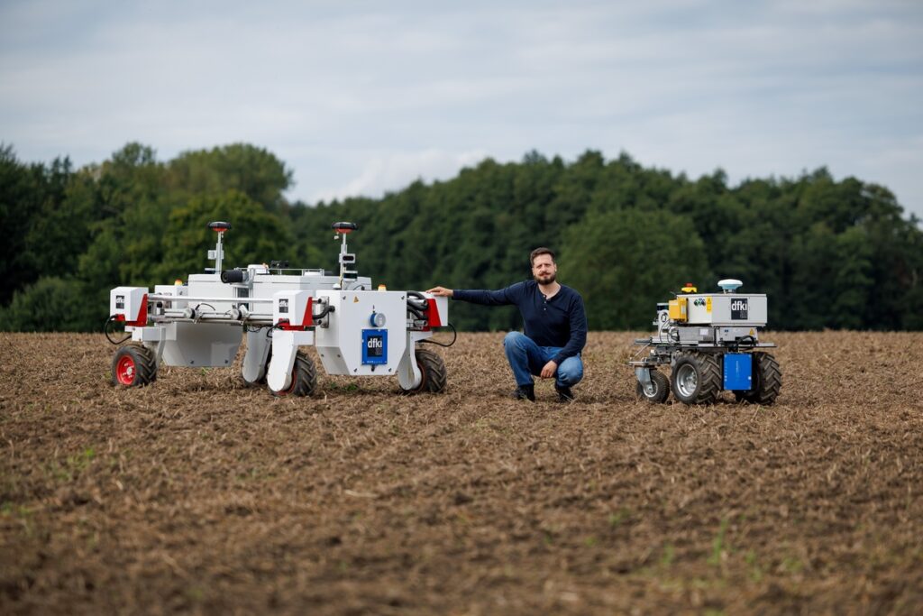 Ein Mann kniet auf einem Feld zwischen zwei weißen Agrarrobotern auf jeweils vier Rädern.