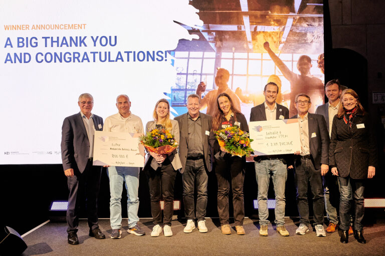 Auf einem Podium lächelt eine Gruppe von neun Menschen in die Kamera, zwei von ihnen halten große Schecks und zwei Blumensträuße.